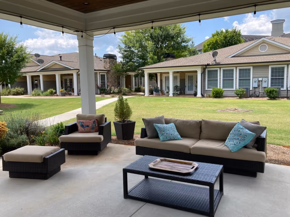 Covered outdoor patio area with cushioned wicker furniture including a sofa with blue patterned pillows, an armchair with a decorative pillow, an ottoman, and a coffee table with a wooden tray. The patio overlooks a well-maintained lawn with shrubs, trees, and single-story residential buildings in the background under a partly cloudy sky.