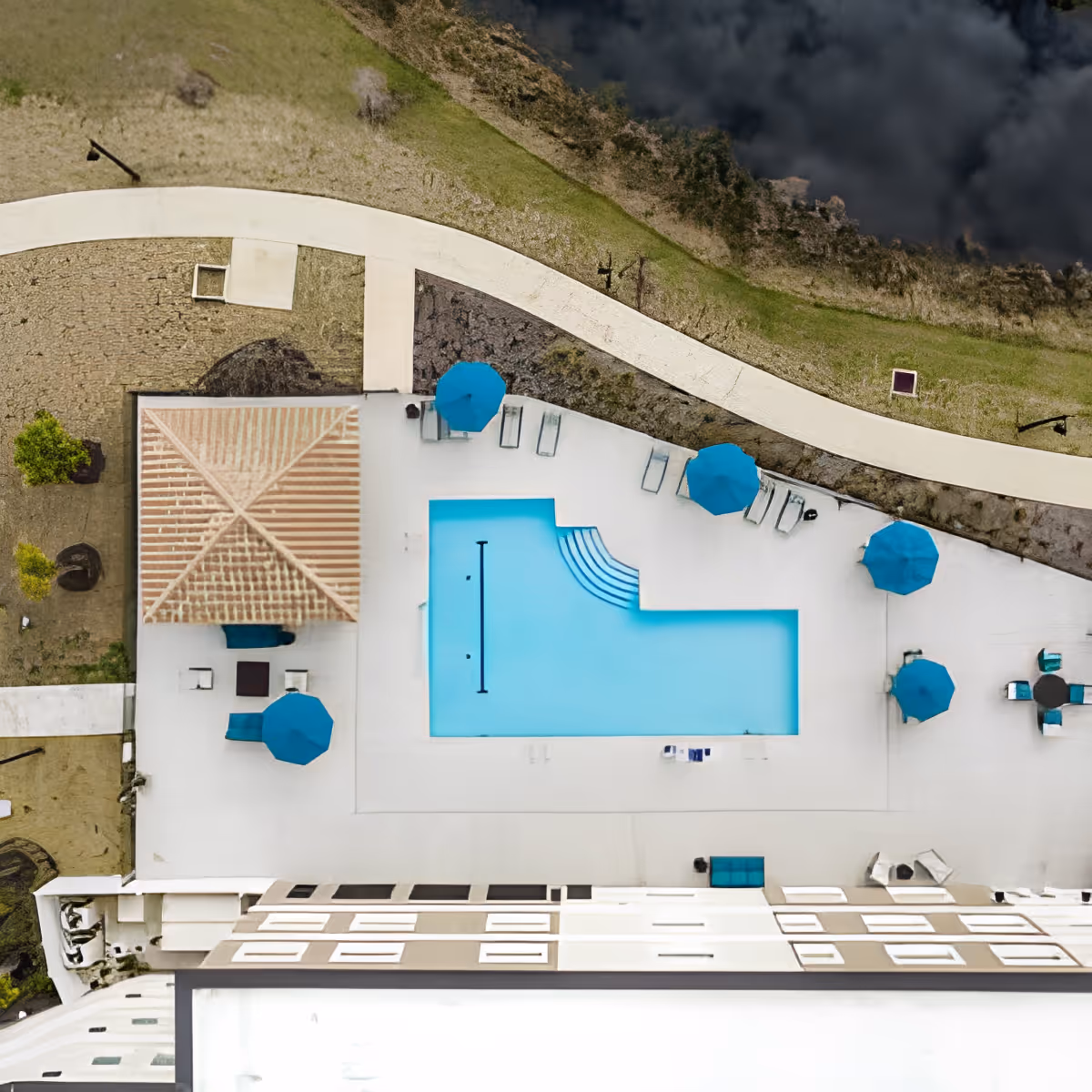 Aerial view of an outdoor swimming pool area with blue umbrellas and lounge chairs surrounding the pool. There is a small covered seating area with a tiled roof on the left side and a curved walkway with grass and landscaping above the pool area.