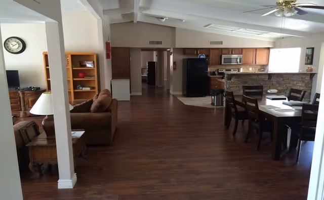 Interior view of a senior living facility showing a spacious open area with wooden flooring. On the left, there is a brown couch, a wooden side table with a lamp, and a bookshelf. In the background, a TV is visible on a wooden stand. On the right side, there is a dining table with chairs and a kitchen area with wooden cabinets, a black refrigerator, a microwave, and a stove. The ceiling has exposed beams and a ceiling fan with lights.