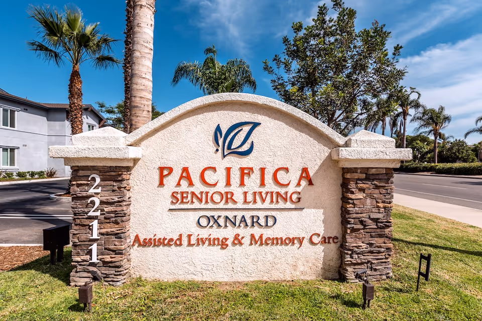 Outdoor view of a large stone and stucco sign for Pacifica Senior Living Oxnard, Assisted Living & Memory Care, with palm trees and a building in the background under a blue sky.