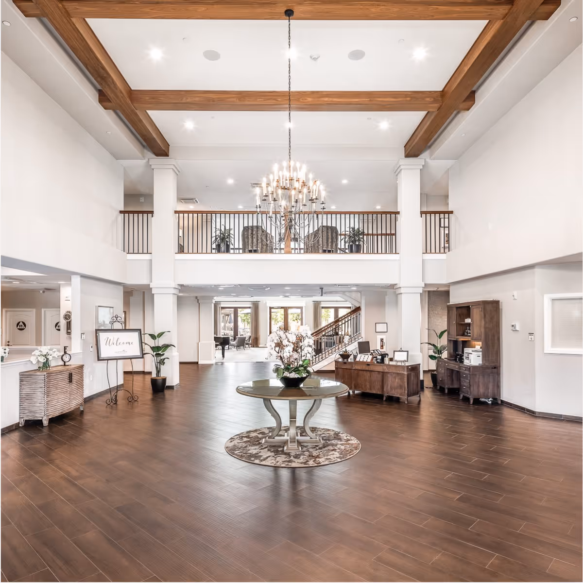 Spacious senior living facility lobby with high ceilings featuring wooden beams, a large chandelier, and a round table with a floral arrangement in the center. The area includes a reception desk, wooden furniture, potted plants, and a welcoming sign. A staircase and seating area are visible in the background.