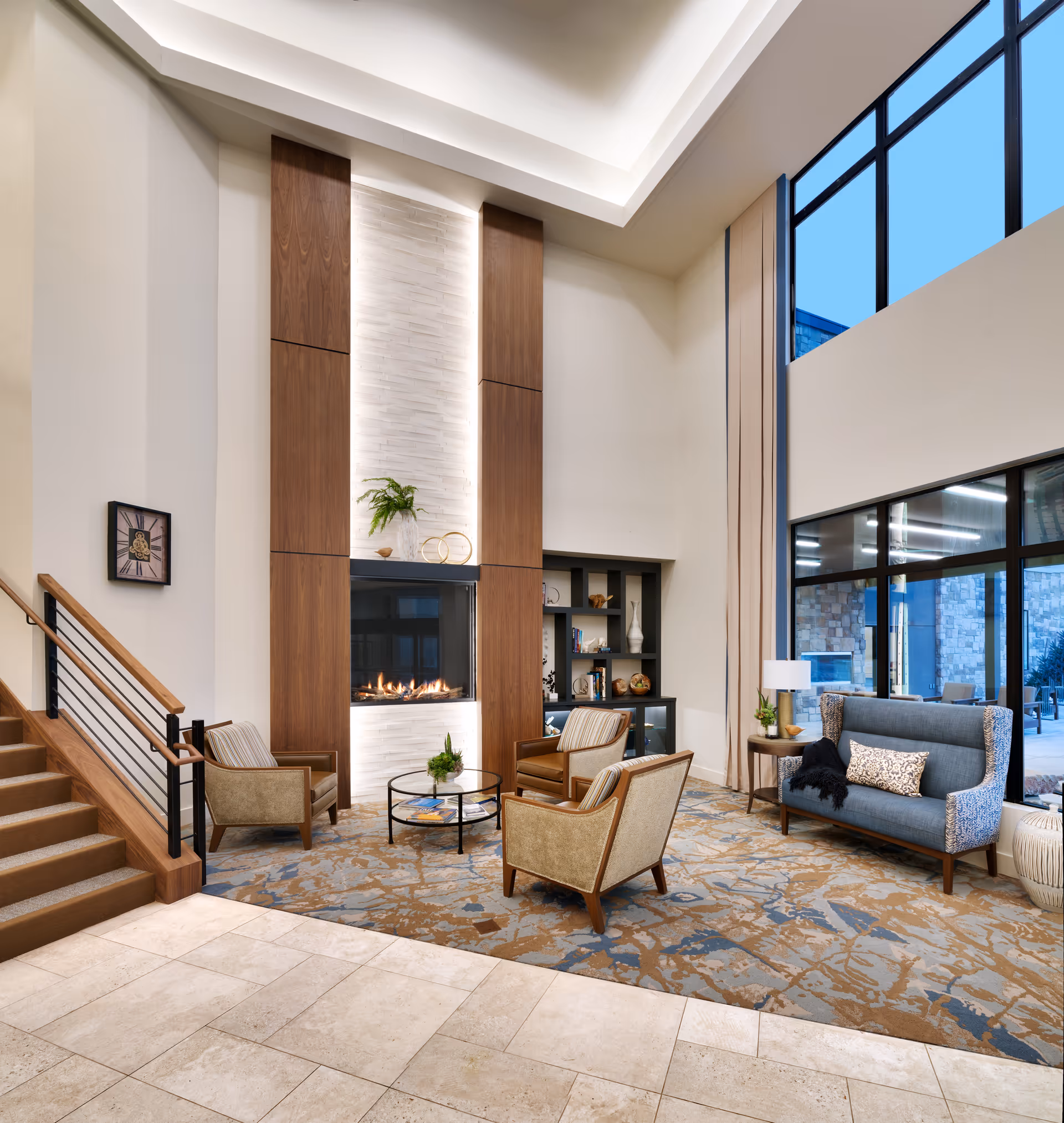 A modern senior living facility lounge area with high ceilings, large windows, and a fireplace. The room features a patterned carpet, three armchairs around a round glass coffee table, a blue cushioned bench with a decorative pillow and throw, and a side table with a lamp and plant. There is a staircase with wooden handrails on the left and built-in shelving with decorative items on the right.