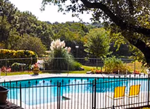 Outdoor swimming pool surrounded by a black metal fence with two yellow chairs placed near the pool. The background features lush green trees and bushes under a clear sky.