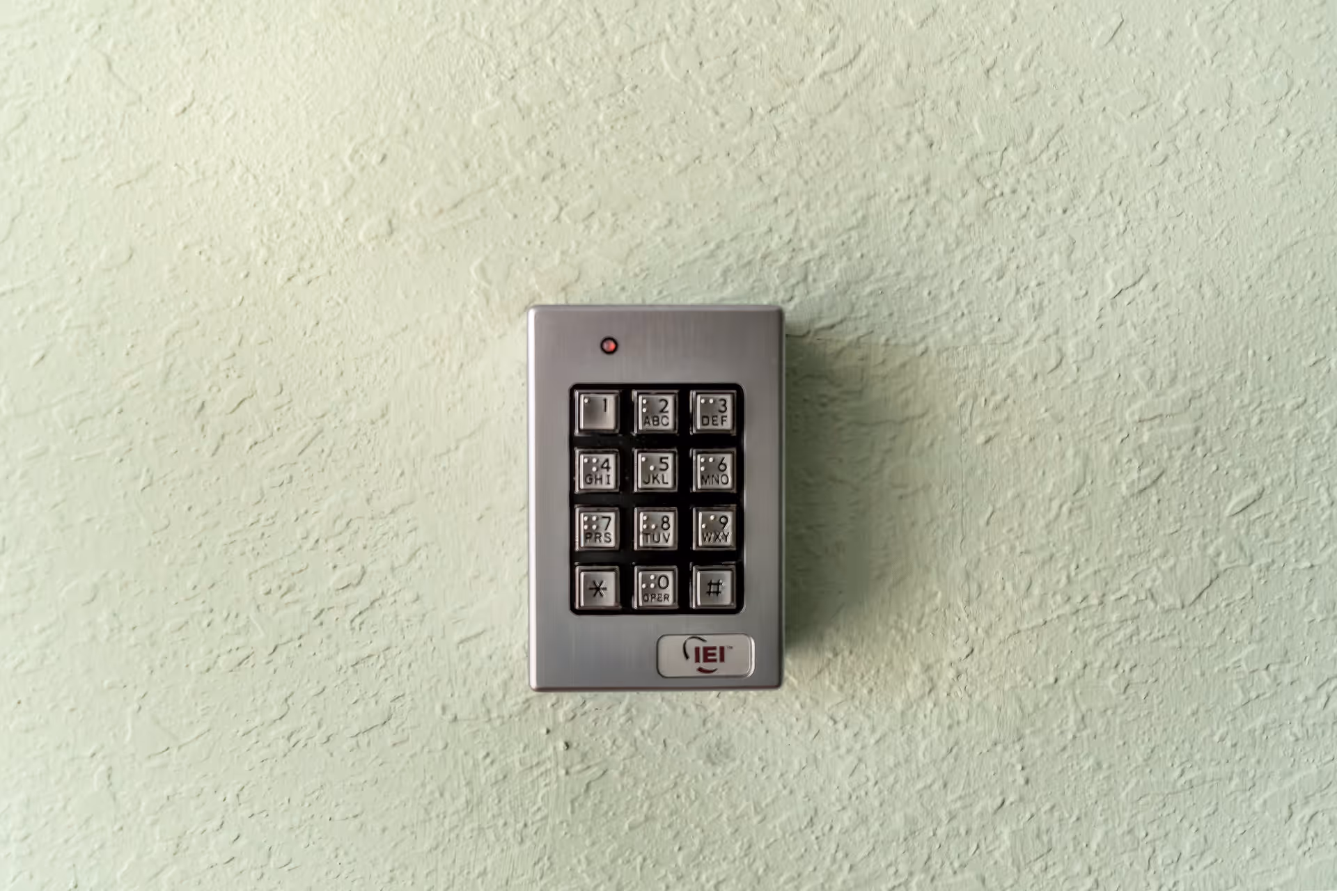 A close-up view of a metallic keypad mounted on a textured light green wall, featuring numeric buttons from 0 to 9 along with star and pound keys, and a small red indicator light above the keypad.