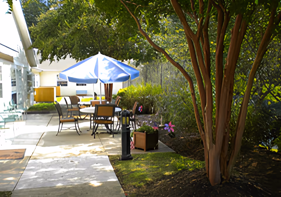 Outdoor patio area with a round table and several chairs under a blue and white umbrella. The patio is adjacent to a building with large windows, surrounded by trees, bushes, and flowering plants.