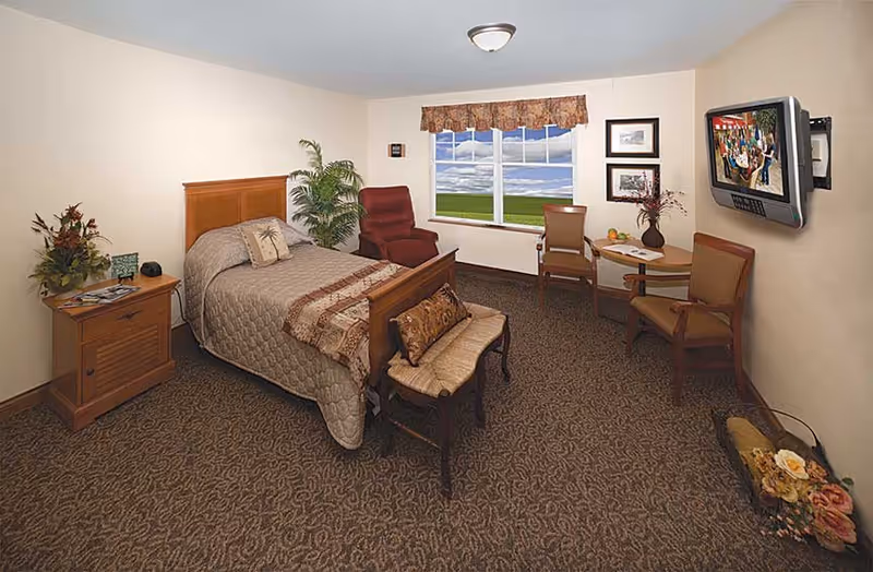 A cozy bedroom in a skilled nursing facility featuring a single bed with a quilted bedspread and decorative pillows. The room includes a wooden nightstand with a plant and magazines, a red armchair, a small table with two chairs near a window showing a scenic outdoor view, framed pictures on the wall, a wall-mounted television, and a decorative bench at the foot of the bed. The carpet has a patterned design and the walls are painted a light color.