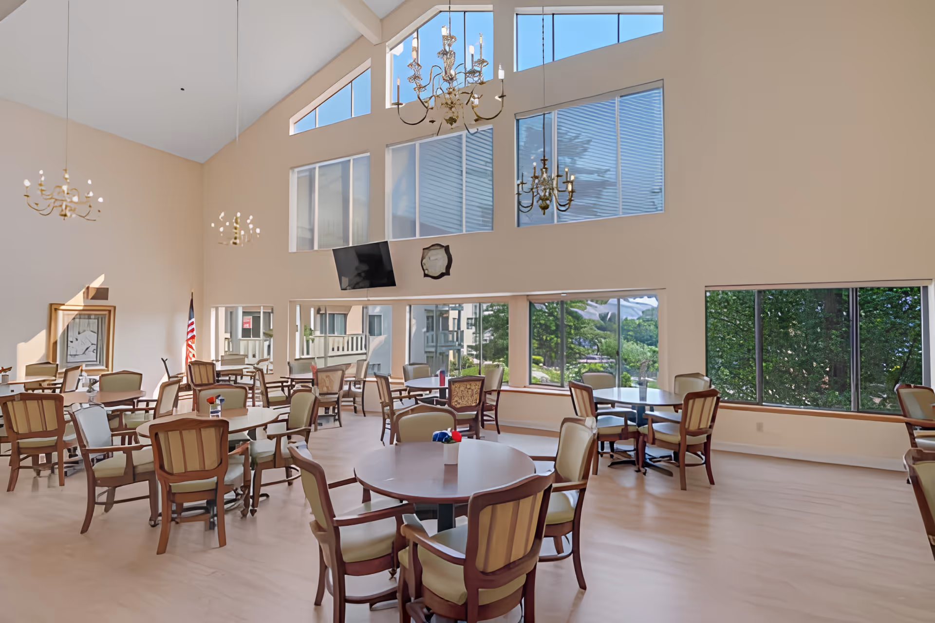 A spacious dining room with multiple round and square tables surrounded by cushioned wooden chairs. The room features large windows letting in natural light and offering views of greenery outside. There are three chandeliers hanging from the high ceiling, and a television mounted on the wall. An American flag is visible in the corner.