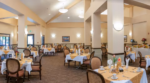 A spacious dining room with multiple round tables covered with white and beige tablecloths, set with cups, plates, and napkins. The room has beige walls, carpeted floors, and several chairs with cushioned backs. There are large windows and doors letting in natural light, and the ceiling has modern light fixtures. A buffet table with flowers and food is visible in the background.