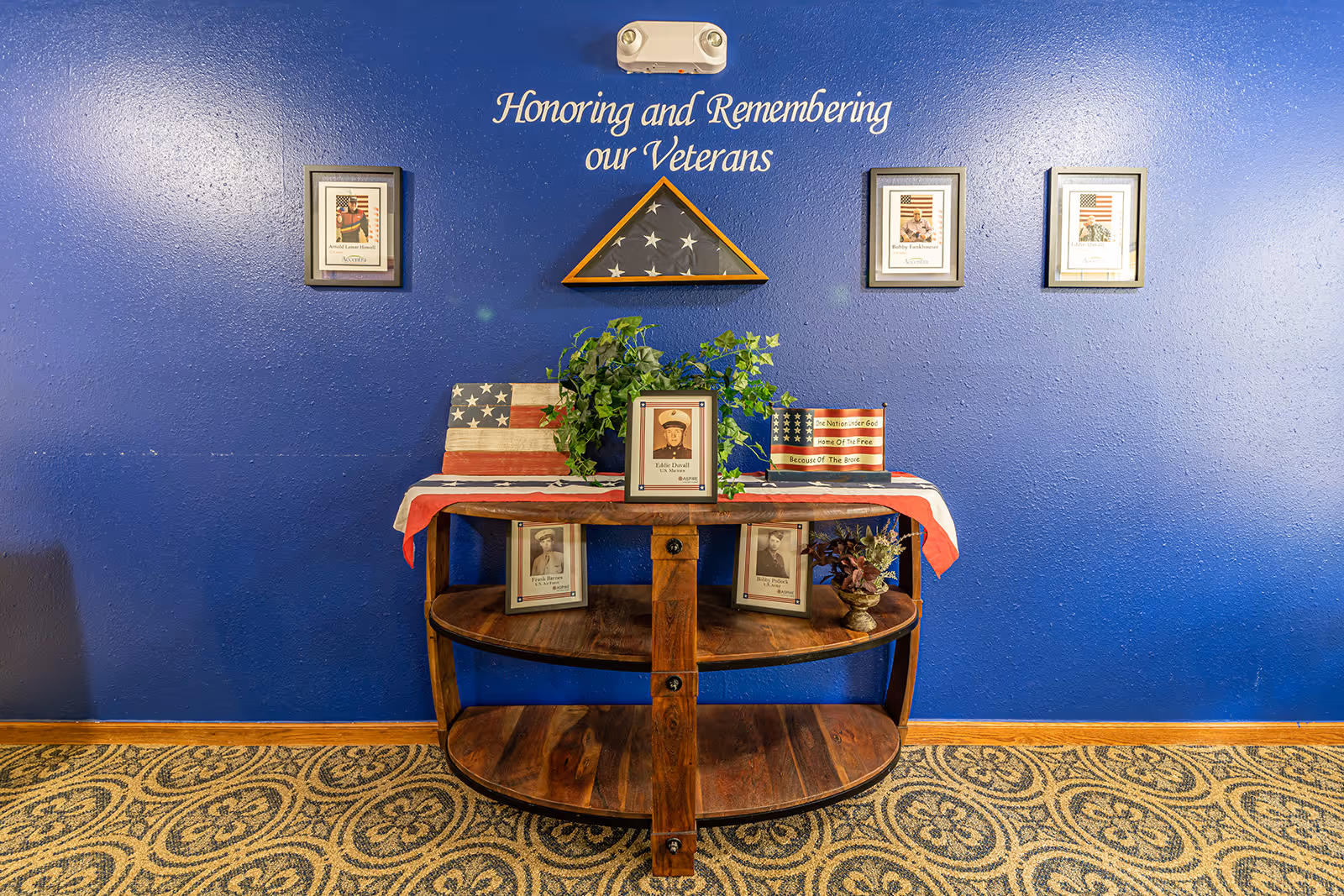 A memorial display honoring veterans with a blue wall background. The wall has the text 'Honoring and Remembering our Veterans' above a folded American flag in a wooden triangular case. Below the flag, there is a wooden table covered with an American flag cloth, framed photos of veterans, a small potted plant, and patriotic decorations. The floor has a patterned carpet.