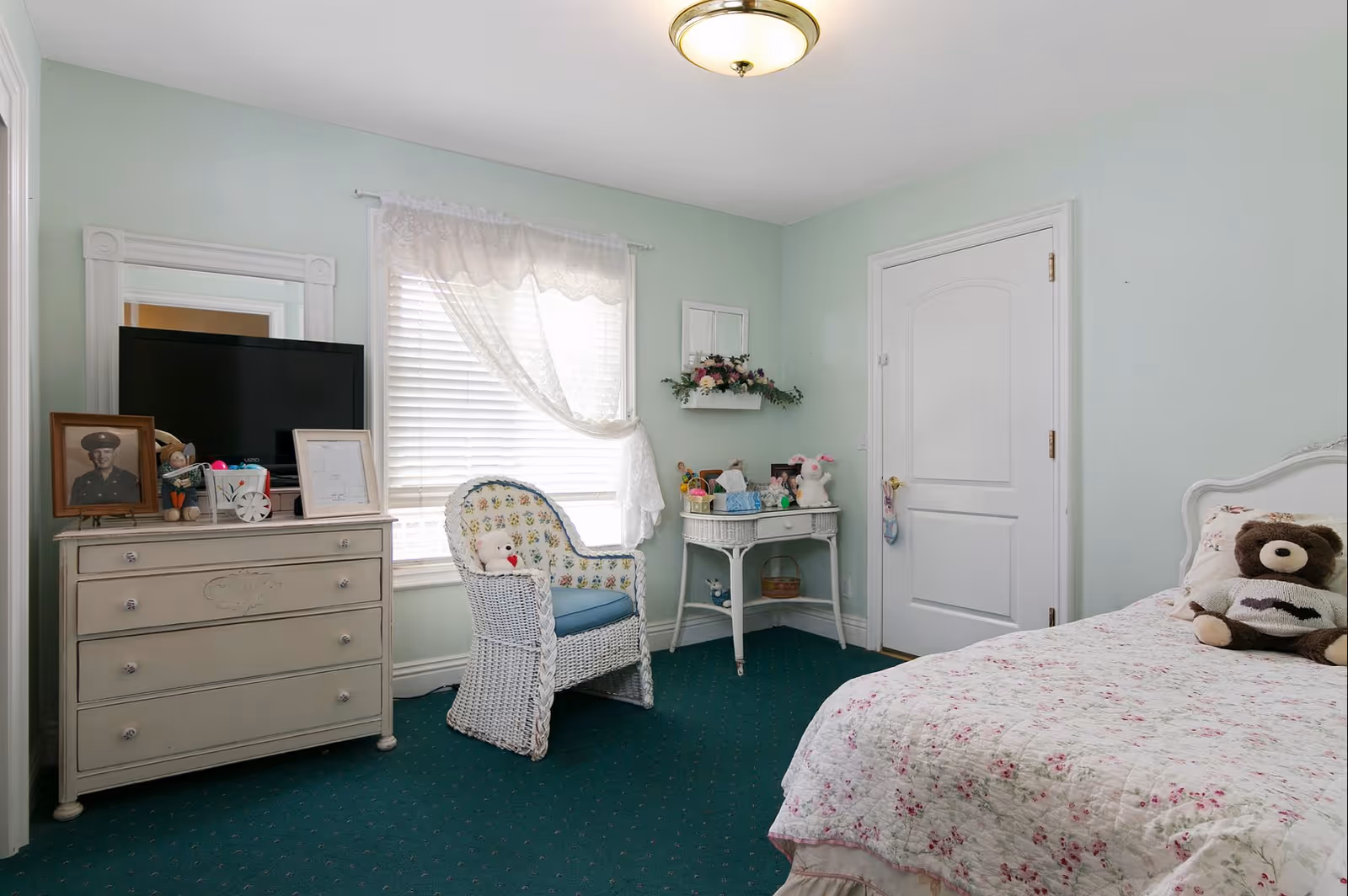 Cozy pastel bedroom with a single bed and stuffed bear, floral quilt, wicker chair, dresser with TV and framed photo, and a small table against a mint-green wall.