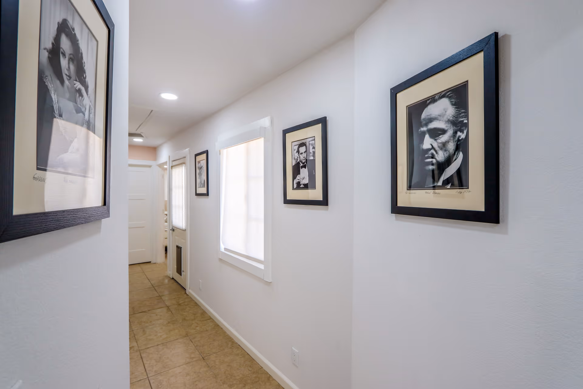 A narrow hallway with beige tiled flooring and white walls, decorated with framed black and white portraits of classic movie stars. There is a window with a closed blind on the right wall and a white door at the end of the hallway.