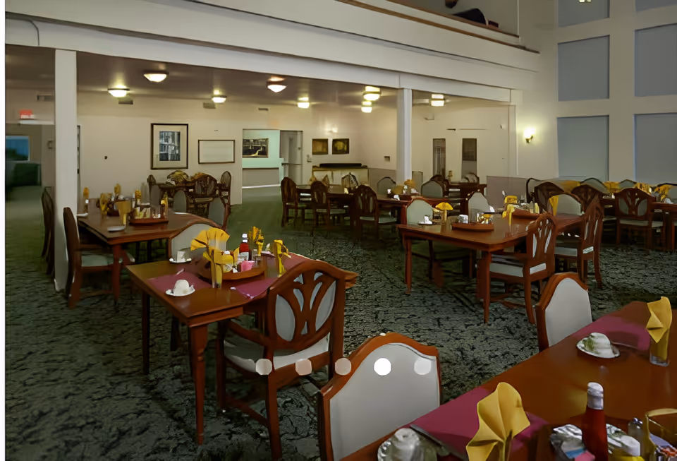 A spacious dining room with multiple wooden tables and chairs arranged neatly. Each table is set with yellow folded napkins, condiments, and tableware. The room has a carpeted floor, white walls, and soft lighting from ceiling fixtures. There are framed pictures on the walls and a high ceiling with a balcony railing visible above.