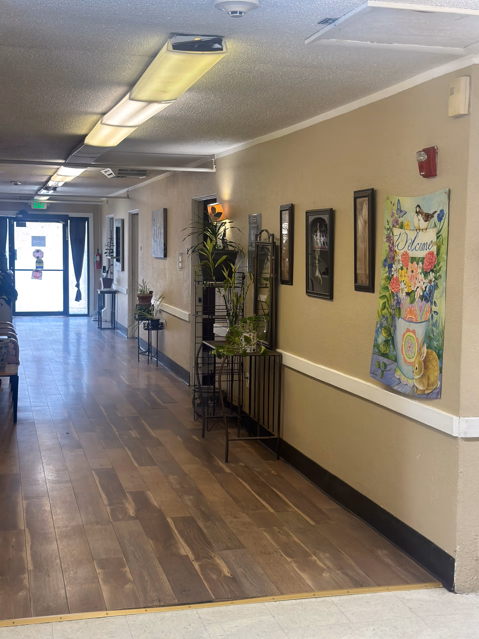 A well-lit hallway in an assisted living facility with wooden flooring, beige walls, and ceiling lights. The right wall is decorated with framed pictures and a colorful 'Welcome' banner featuring flowers, a bird, and a rabbit. There are several potted plants on metal stands along the hallway. At the end of the hallway, there is a glass door with an exit sign above it.
