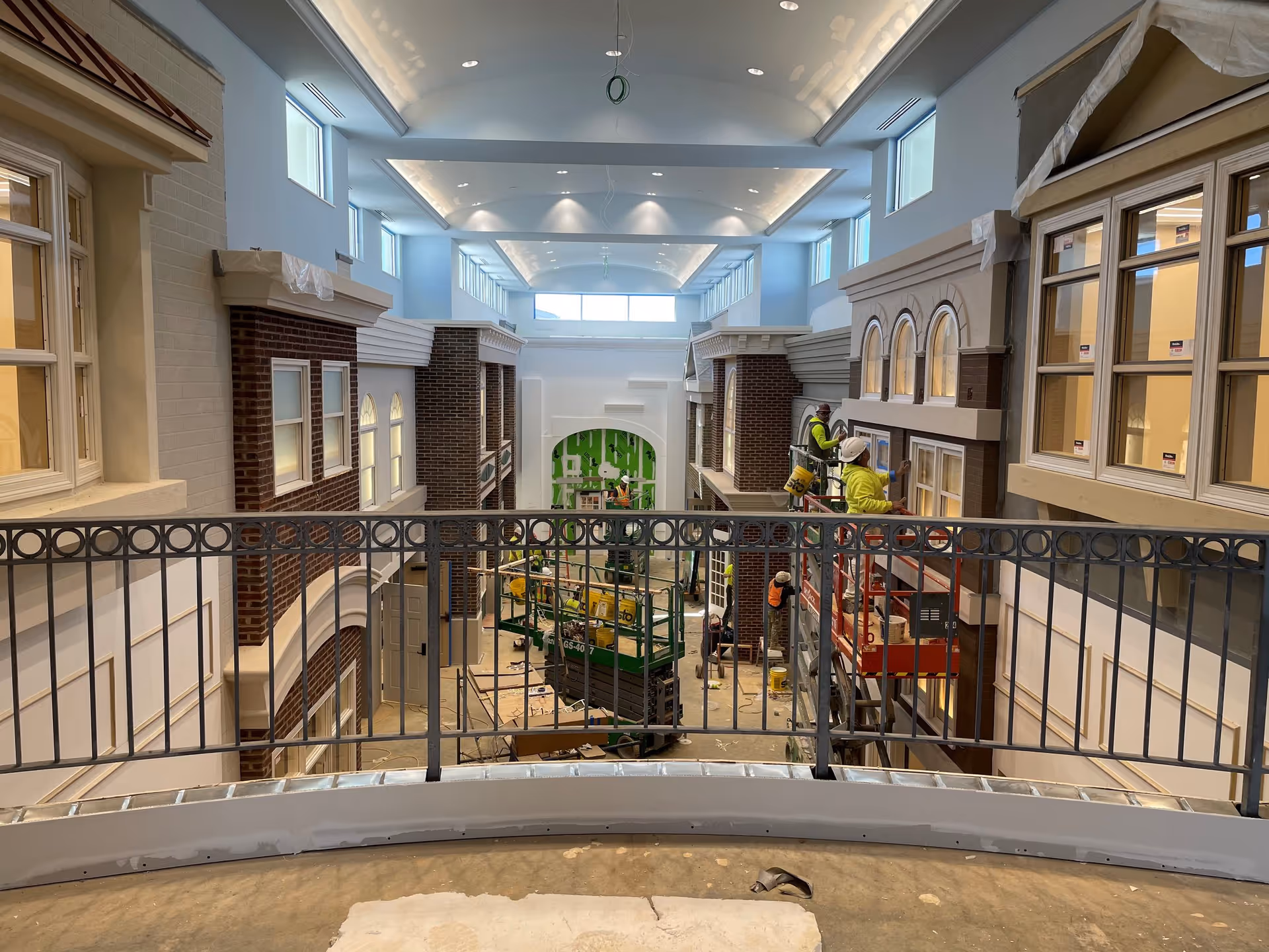 Interior view of a large, high-ceilinged space under construction with workers on lifts installing windows and finishing walls. The area features architectural elements resembling a street with brick and stone facades, multiple windows, and a metal railing in the foreground.