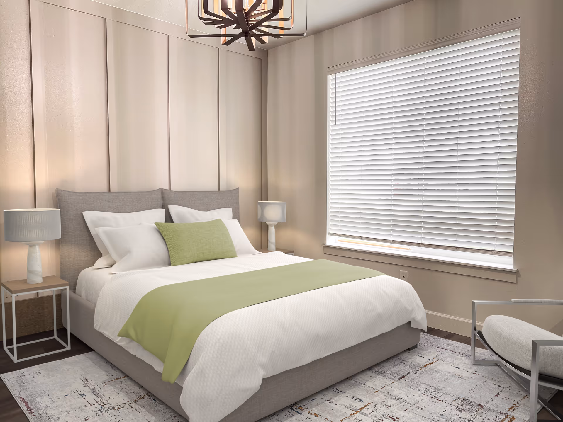 A modern bedroom with a large bed featuring white bedding and a light green throw and pillow. The room has two bedside tables with matching lamps, a large window with white blinds, a patterned area rug, and a contemporary armchair with a metal frame. A unique chandelier hangs from the ceiling.