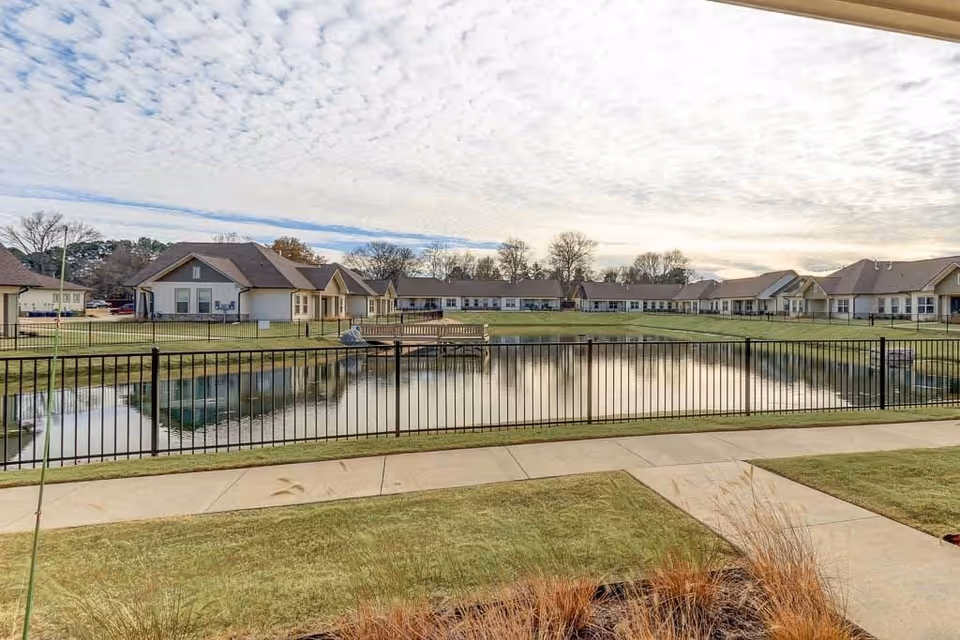 View of a senior living facility with single-story buildings surrounding a pond. There is a black metal fence around the pond and a sidewalk in the foreground. The sky is partly cloudy.