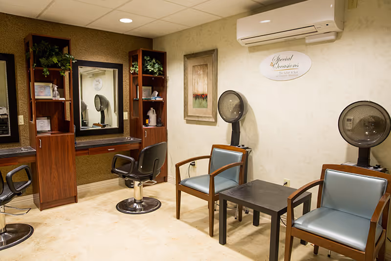 Interior view of a salon area in a retirement community featuring two black salon chairs in front of mirrors and wooden shelving units with plants and hair products. There are also two blue cushioned chairs with wooden arms next to a small black table, and two hair dryers mounted on stands against the wall. A sign on the wall reads 'Special Occasions Hair Salon & Spa at Country Meadows.'