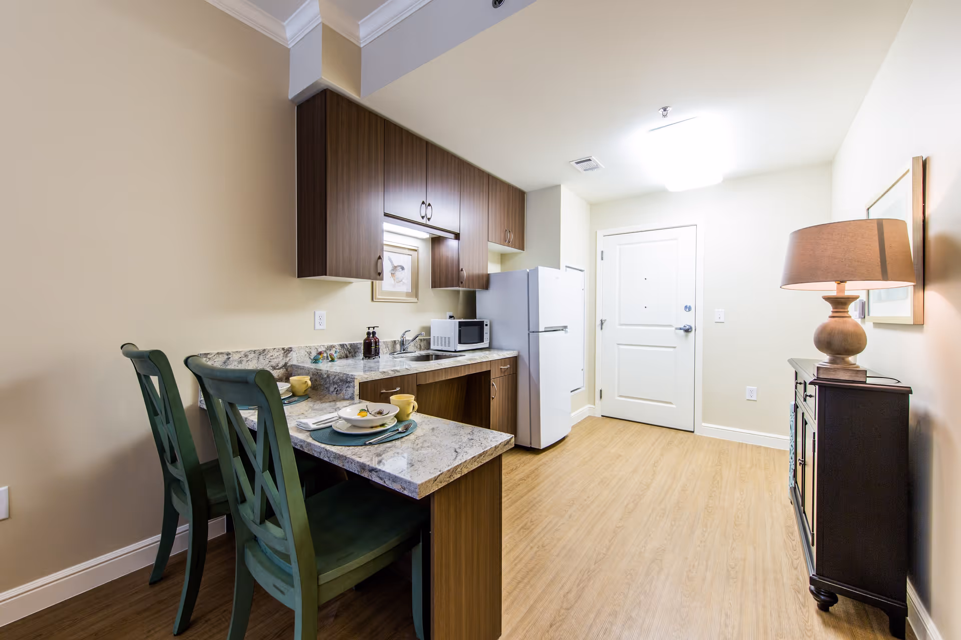 A small kitchen area with wooden cabinets, a white refrigerator, a microwave, and a granite countertop with two green chairs. The kitchen is adjacent to a hallway with a white door and a wooden floor. A lamp on a dark wooden cabinet is visible on the right side.