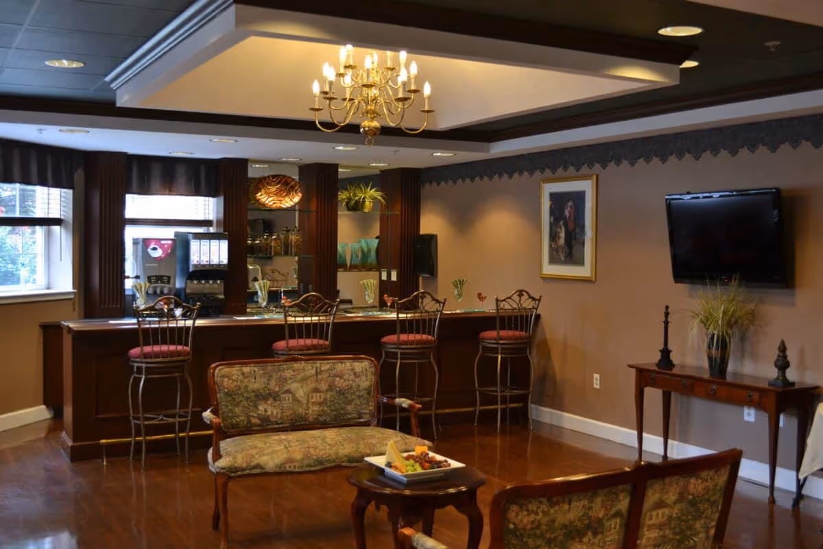 Cozy senior living common room with a small bar, barstools, seating area, chandelier and a wall-mounted TV.