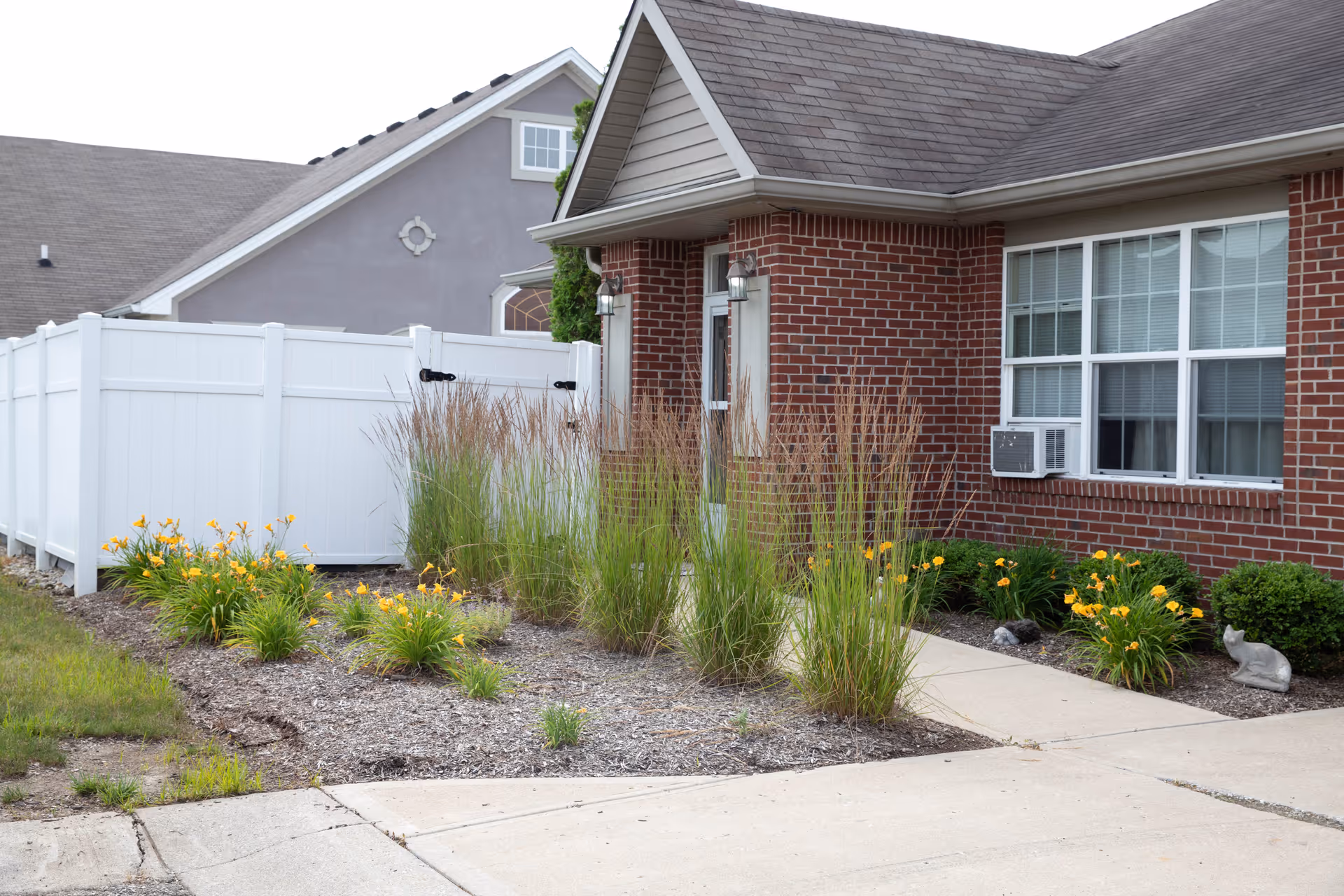 Exterior view of a brick building with a window air conditioning unit, a white fence, and a landscaped garden featuring tall grasses and yellow flowers along a concrete walkway.