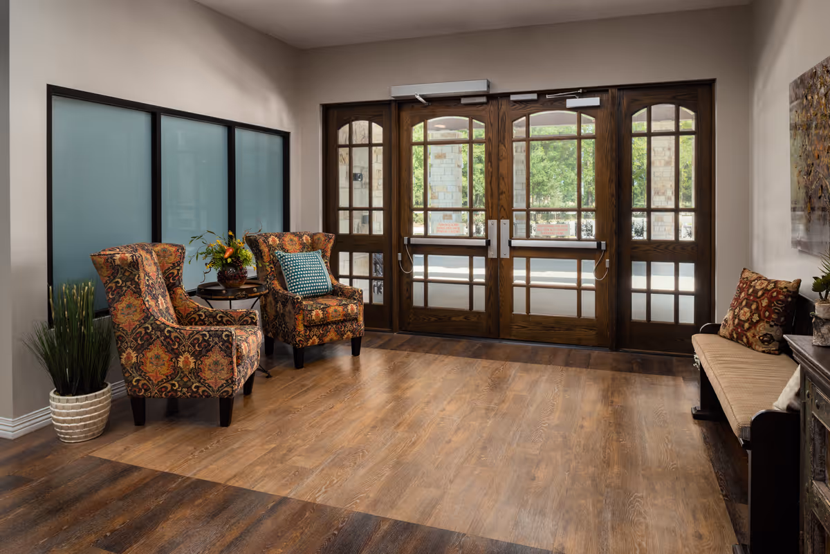 A cozy senior living facility entrance area with two patterned armchairs and a small round table with a flower arrangement between them on the left, a wooden bench with cushions on the right, and large wooden double doors with glass panels letting in natural light.