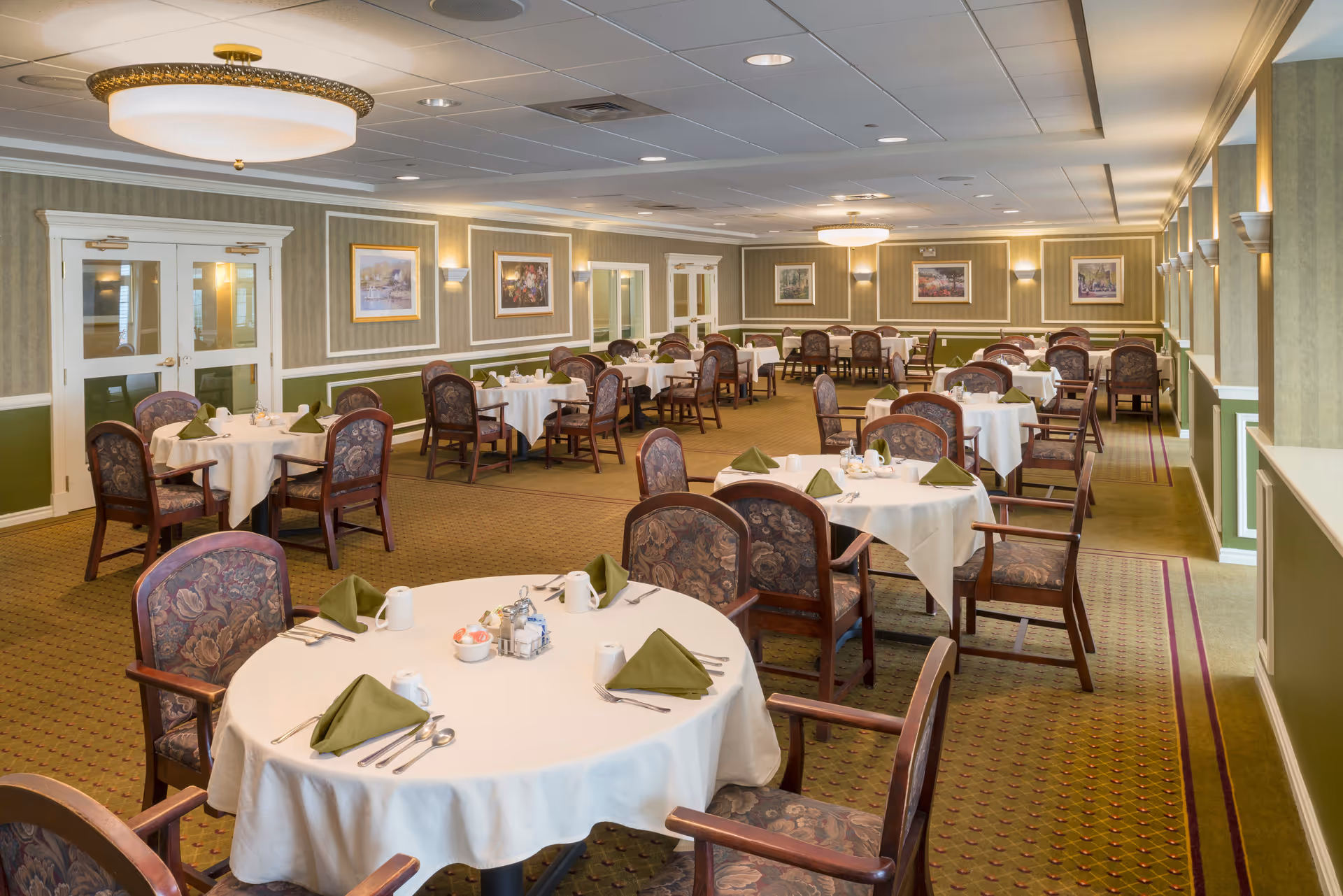 A spacious dining room with multiple round tables covered with white tablecloths, each set with green folded napkins, cups, and silverware. The room has patterned carpet, upholstered chairs, framed artwork on the walls, and soft lighting from ceiling fixtures and wall sconces.