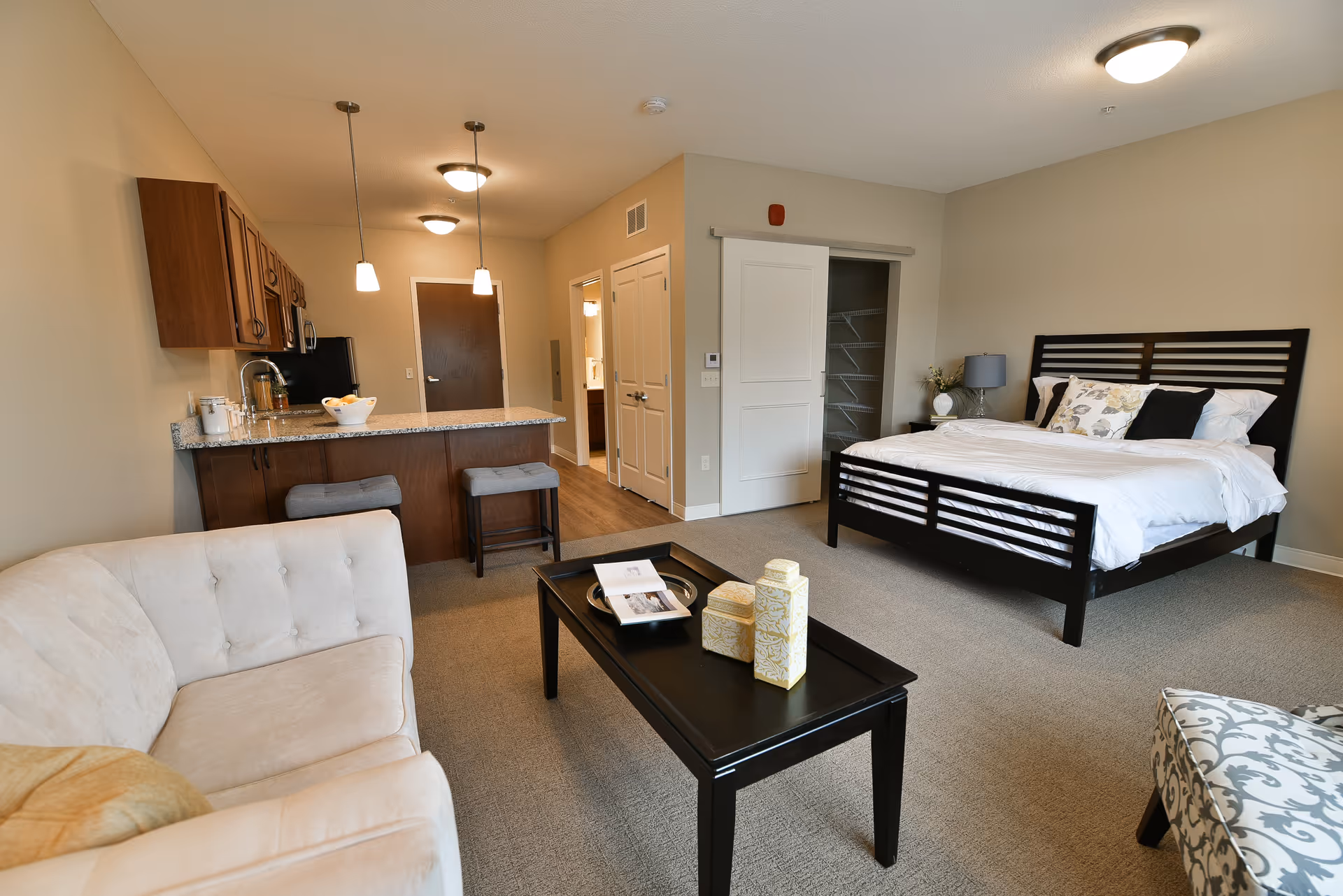 A studio apartment interior at Havenwood of Richfield featuring a bed with white bedding and decorative pillows, a black coffee table with decorative items and an open book, a beige sofa, a patterned armchair, and a kitchen area with wooden cabinets, granite countertop, and two stools. The room has beige walls and carpeted flooring, with a closet and a bathroom door visible in the background.