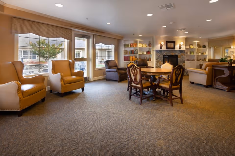 A cozy living room area in a senior care facility with several armchairs and a round wooden table with four chairs. The room features large windows with views of an outdoor courtyard, built-in bookshelves with decorative items, a stone fireplace with wall sconces, and soft lighting creating a warm atmosphere.
