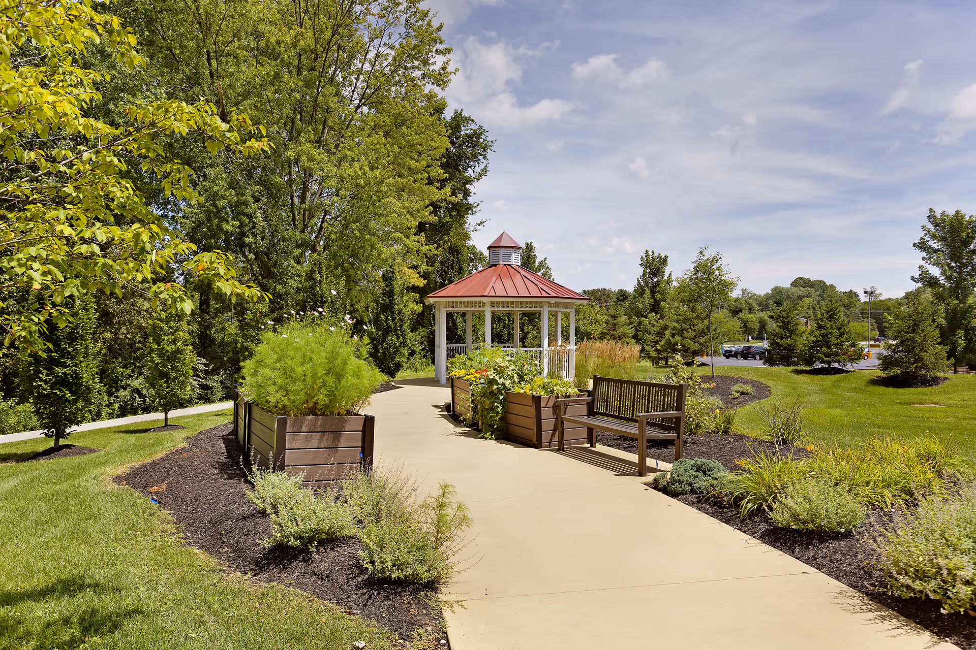 A paved walkway in a garden area with green trees and shrubs on both sides, leading to a white gazebo with a red roof. There are wooden benches and planter boxes with plants along the path under a partly cloudy sky.