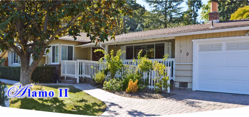 Single-story residential building with a white fence and a garden in front, featuring a tree, shrubs, and a paved walkway leading to the entrance. The house has beige siding, a white garage door, and large windows with curtains.