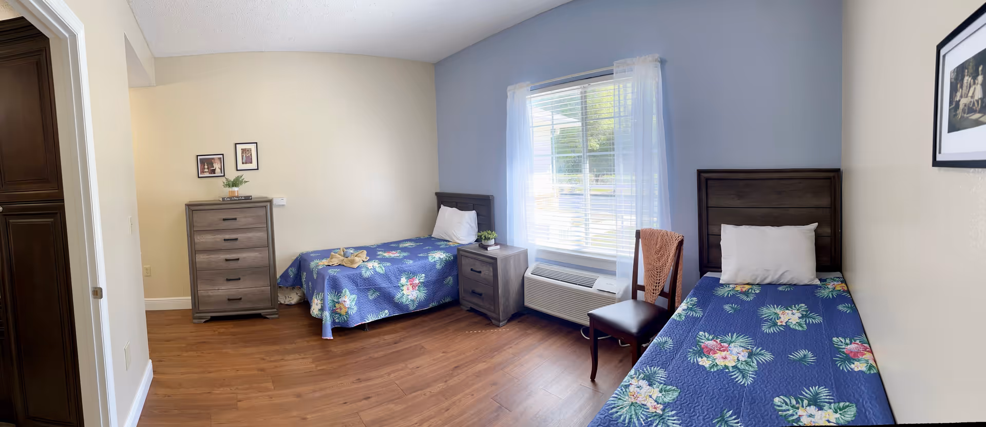 Bright bedroom with two single beds, a dresser and nightstand, a window with blinds, and wood flooring.