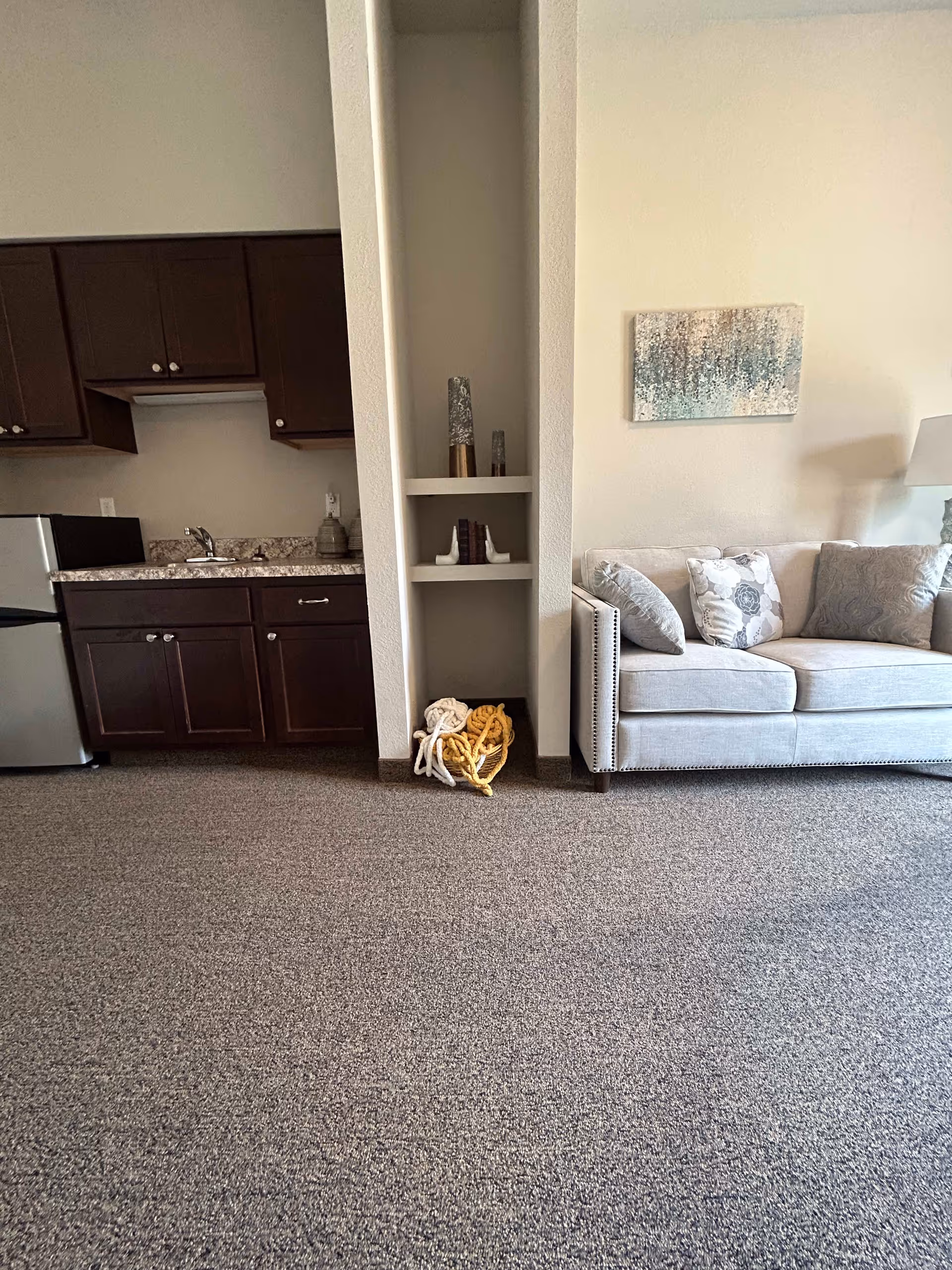 Open studio interior showing a beige sofa with decorative pillows, built-in shelving, and a small kitchenette with dark cabinets and a mini fridge.