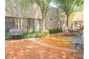 Outdoor patio area with brick flooring, surrounded by trees and greenery. There are metal benches and a table with chairs arranged for seating in a shaded courtyard next to a building.
