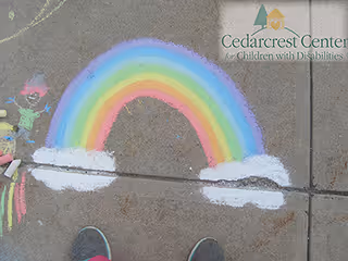 A colorful sidewalk chalk rainbow with clouds drawn on concrete and the Cedarcrest Center logo in the top-right corner.
