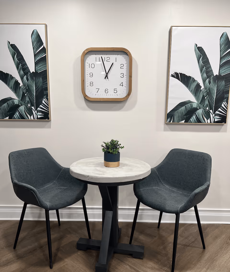 A small round table with a potted plant on top, flanked by two dark green upholstered chairs with black legs. Above the table is a square wall clock with a wooden frame showing the time as 11:55. On either side of the clock are framed botanical prints featuring large green leaves. The setting has a light-colored wall and wood-patterned flooring.