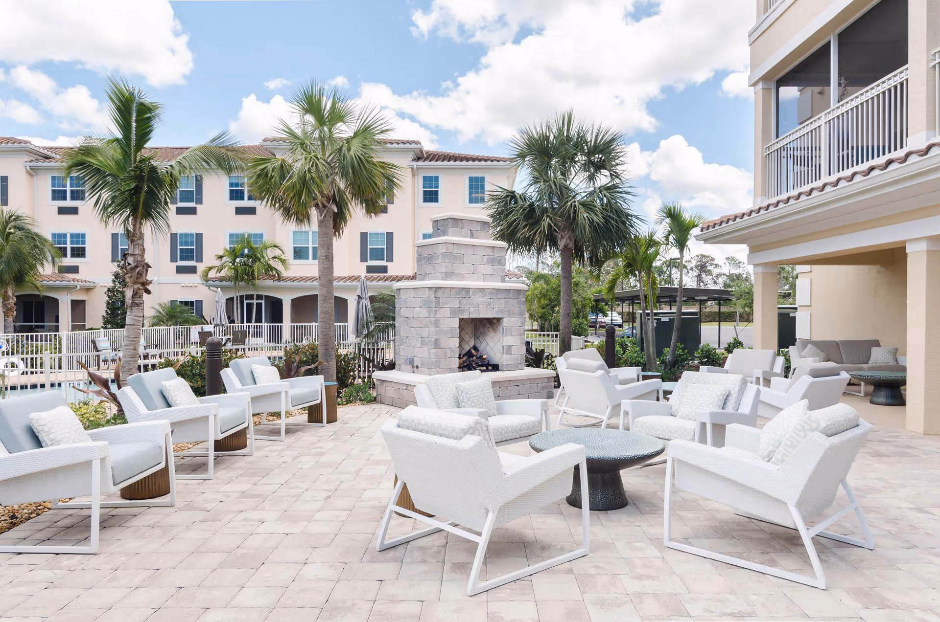 Outdoor patio area with white cushioned chairs arranged around a central round table and a stone fireplace. Palm trees and a multi-story building with balconies are visible in the background under a partly cloudy sky.