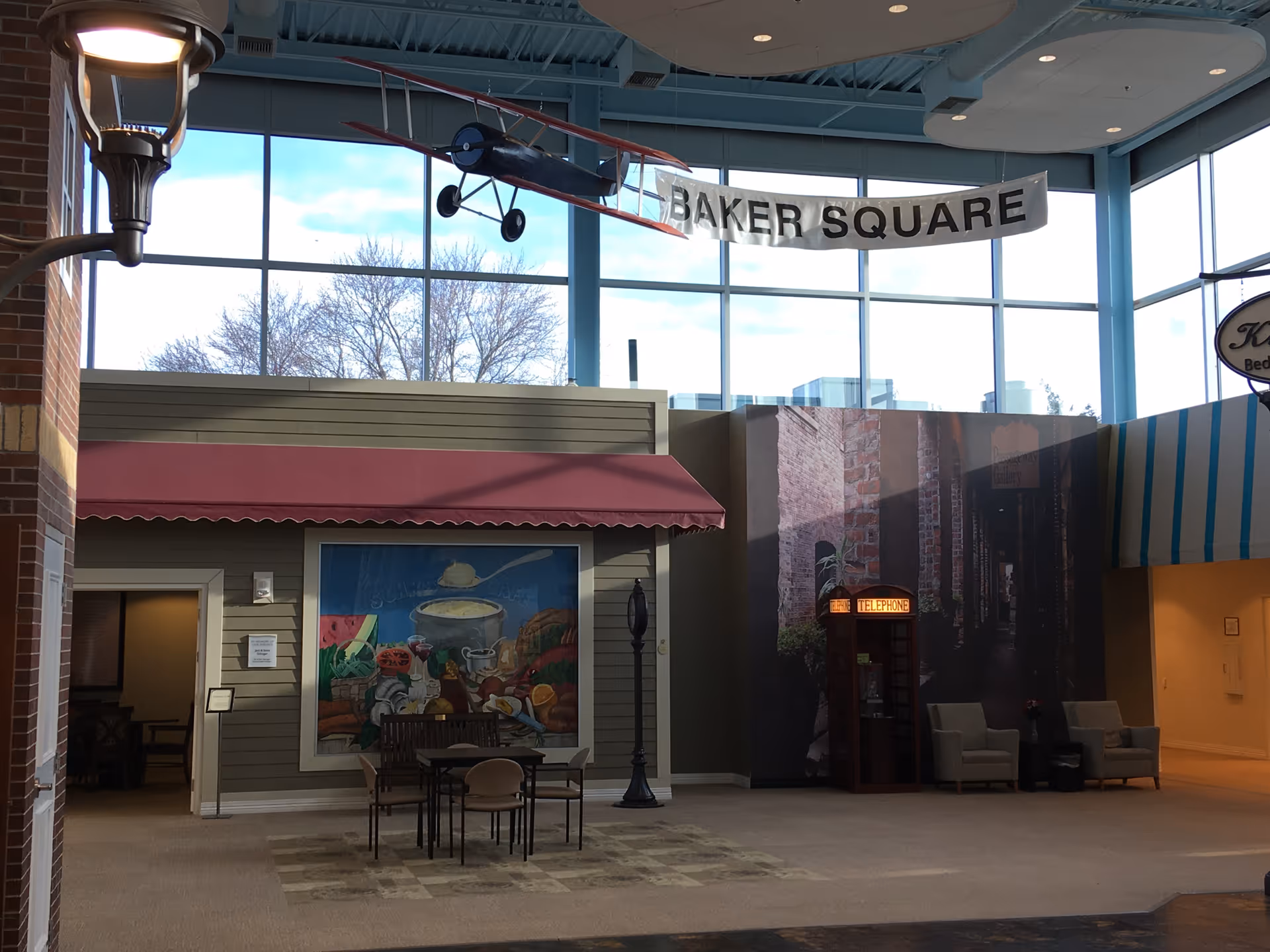 Indoor common area with mock storefronts, tables and chairs, a hanging model airplane and a banner reading 'BAKER SQUARE'.