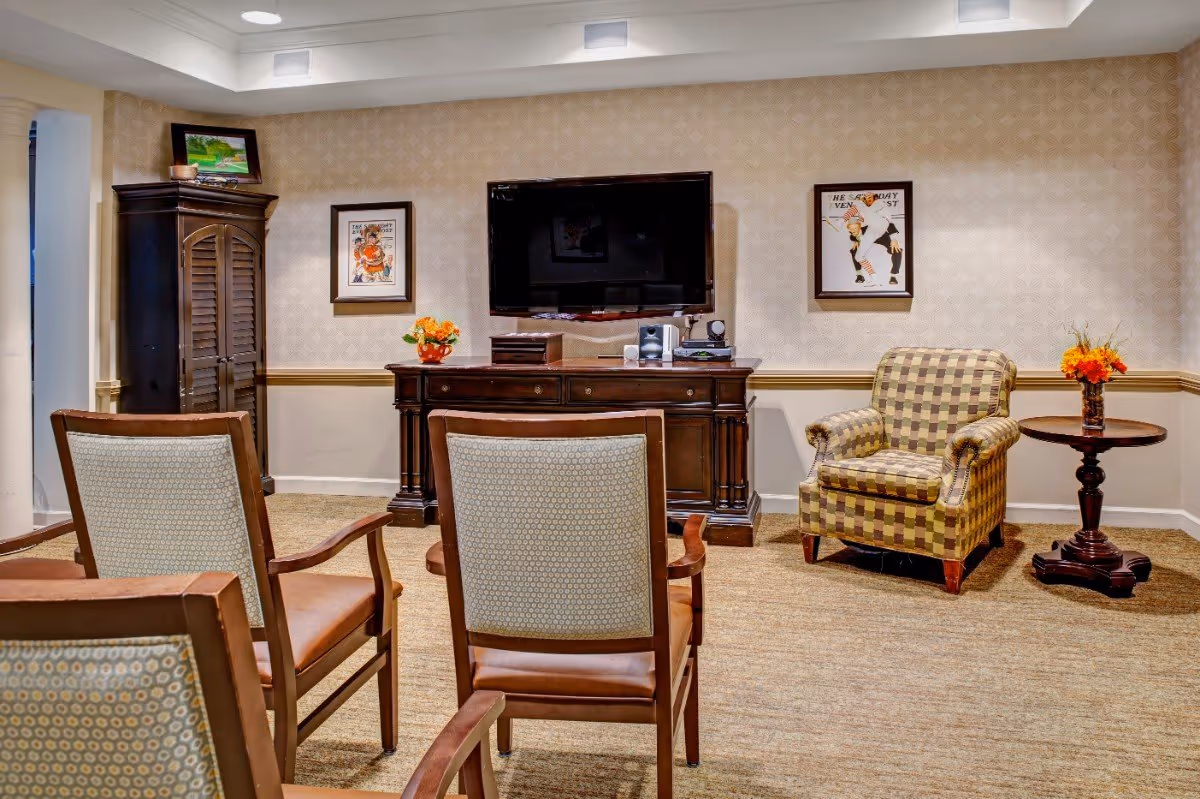 A cozy sitting area in a senior living facility with four wooden chairs facing a wall-mounted flat screen TV. The room features a patterned armchair next to a small round wooden table with a vase of orange flowers. There is a dark wooden cabinet beneath the TV and framed artwork on the wallpapered wall.