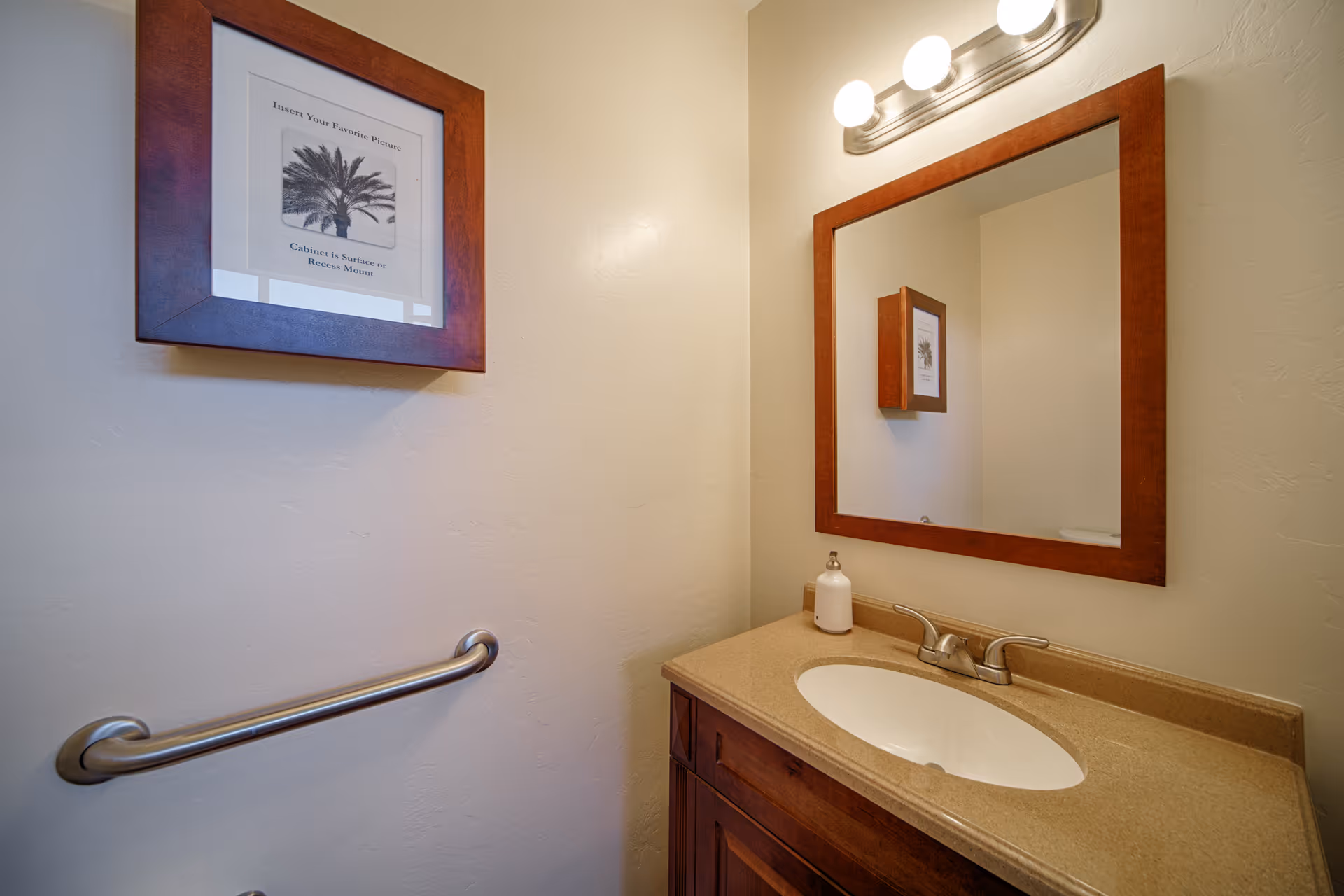 A small bathroom with a beige countertop and an oval sink. Above the sink is a rectangular mirror with a wooden frame and a light fixture with two bulbs. On the wall to the left, there is a framed picture of a palm tree and a metal grab bar below it.