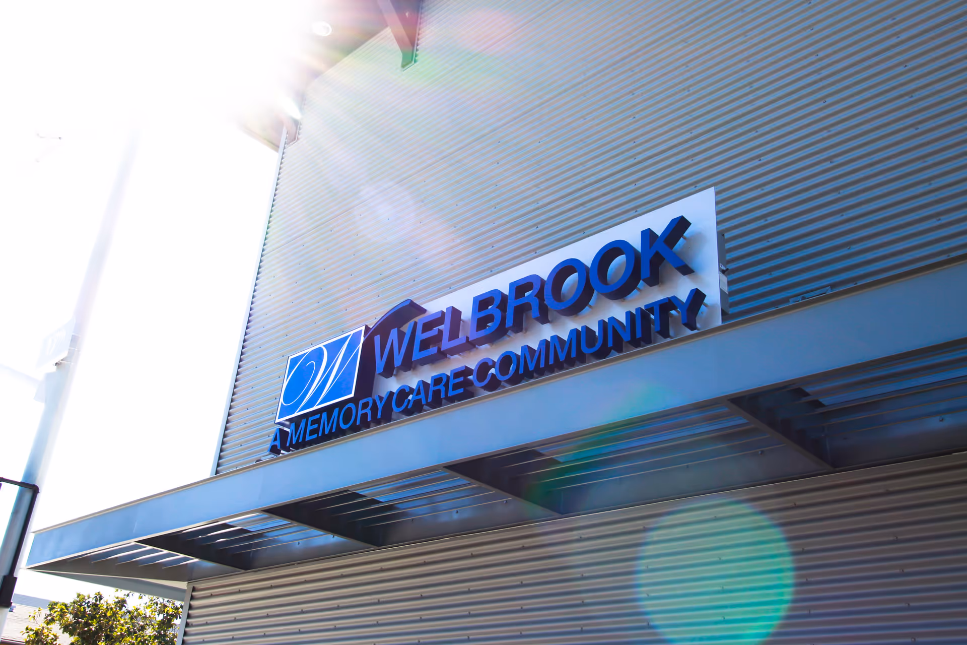 Exterior view of the Welbrook Memory Care Community building with a metal facade and a sign displaying the facility's name. Sunlight creates lens flare in the image.