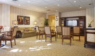 A spacious living room area with several wooden chairs arranged in rows facing a large wooden entertainment center with a television. There is a beige sofa against the wall with two framed artworks above it, two table lamps on side tables, a potted plant in the corner, and a clock on the wall. The room has carpeted flooring and a ceiling with recessed lighting.