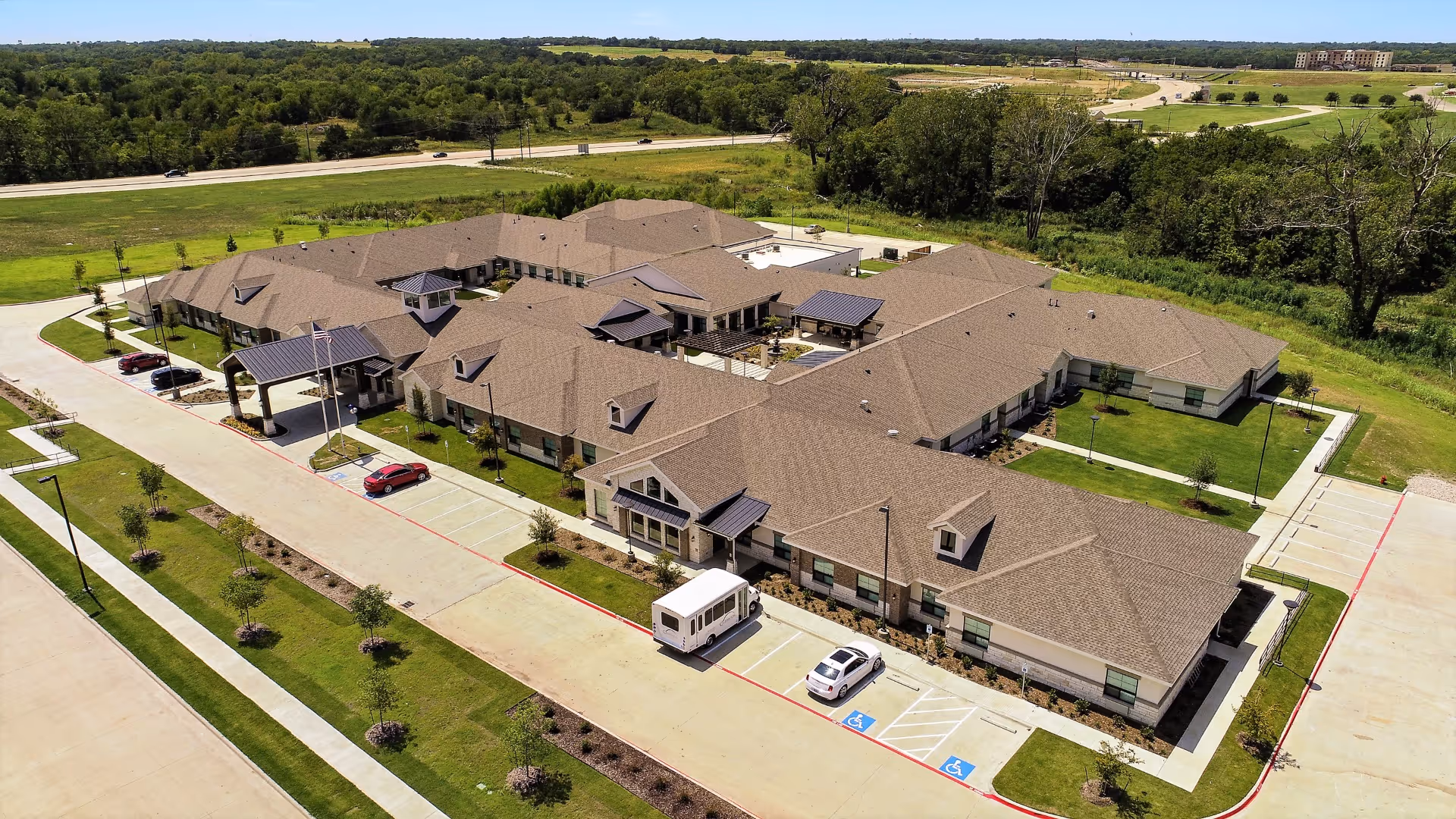 Aerial view of Celebration Senior Living of Denison, showing a large single-story building with multiple wings, surrounded by green lawns and parking spaces including handicapped spots. The building has a brown roof and beige walls, with a covered entrance and several small trees planted around the property. A road and forested area are visible in the background under a clear blue sky.