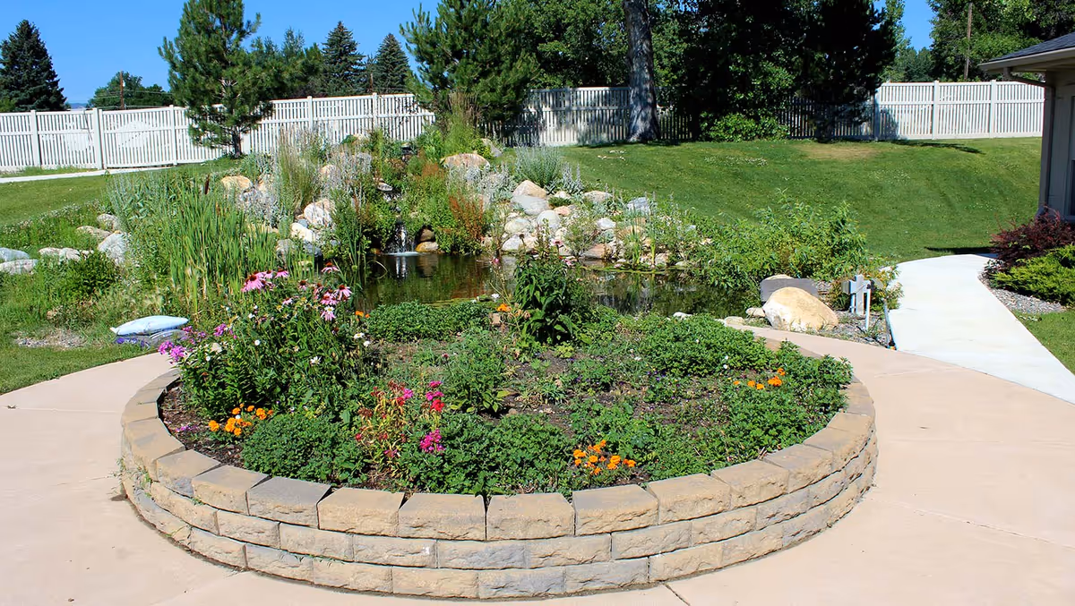 A landscaped outdoor garden area with a circular raised flower bed filled with various colorful flowers and greenery. Behind the flower bed is a small pond with rocks and plants surrounding it. There is a white wooden fence in the background and a concrete pathway curving around the garden area. Trees and a building are visible on the right side.