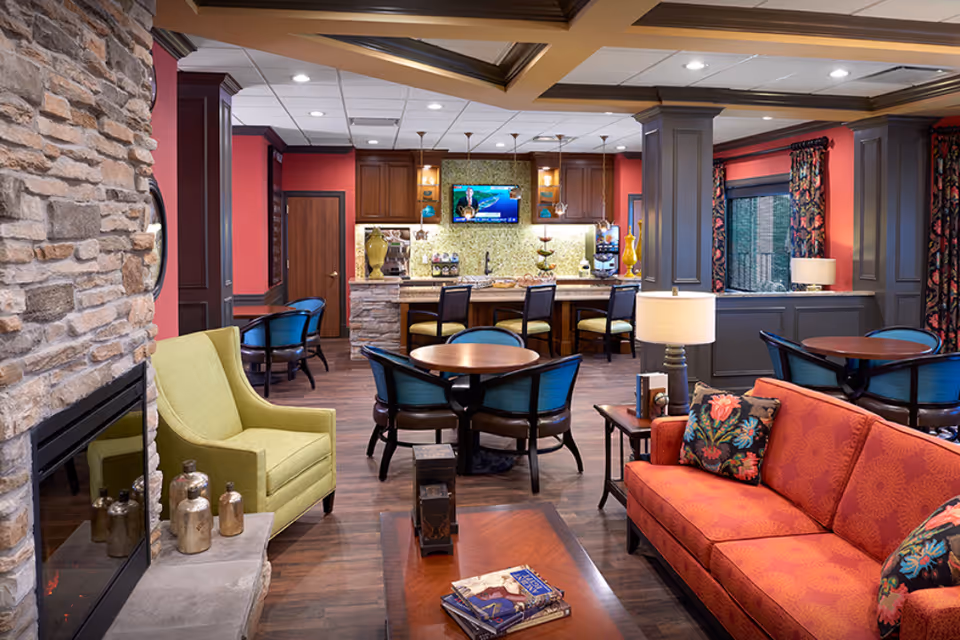 A cozy and well-decorated common area in an assisted living facility featuring a stone fireplace, a green armchair, a red sofa with floral cushions, round wooden tables with blue chairs, a countertop bar with high chairs, and a wall-mounted TV. The room has warm lighting, wooden flooring, and red and gray walls with floral curtains.