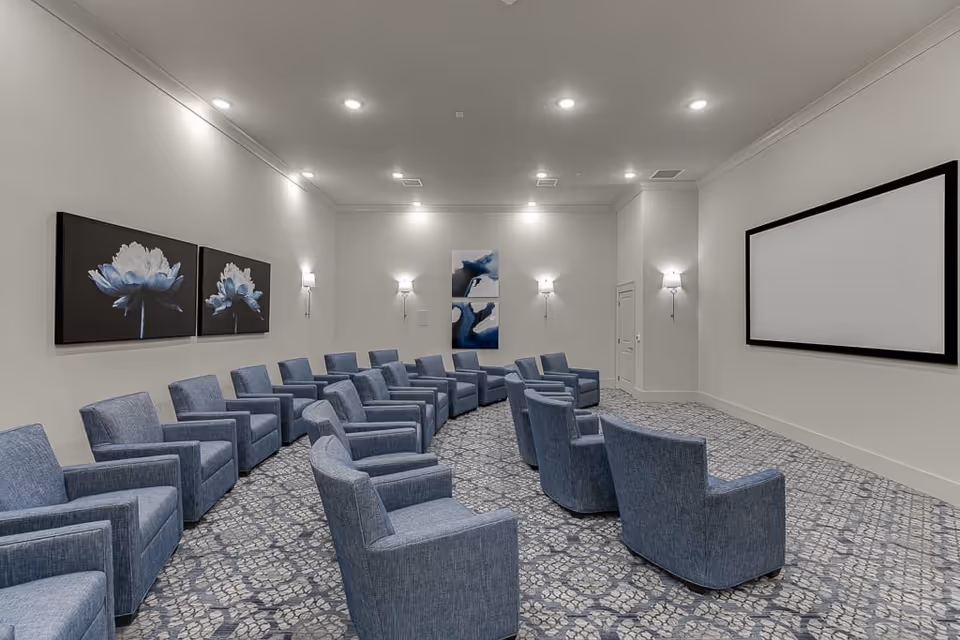 Interior screening room with rows of blue armchairs facing a large projection screen and floral artwork on the walls.