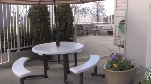 Outdoor patio area with a round white table and curved benches under an umbrella. There are tall green plants along a white fence, a large potted plant with flowers, and a bench in the background near a railing.