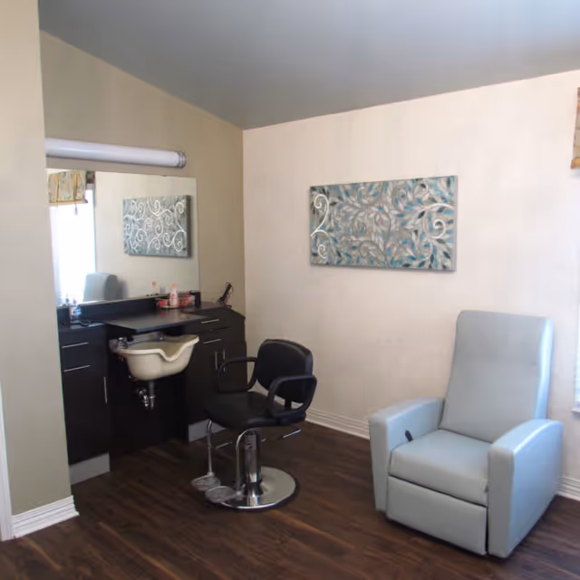 Small salon-style room with a styling chair and wash sink, a large mirror, and a light blue recliner on wood flooring.