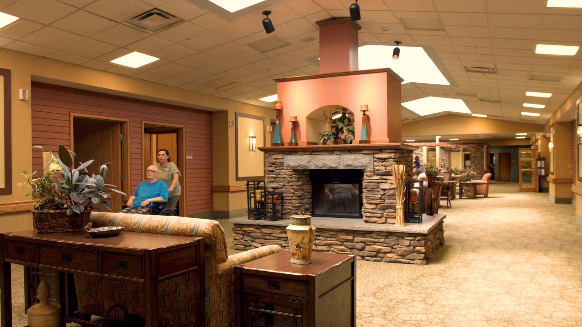 A spacious and warmly lit interior common area of a senior living facility featuring a stone fireplace in the center, comfortable seating with patterned upholstery, wooden tables with decorative items, and a woman pushing an elderly man in a wheelchair along a hallway with doors and wall sconces.