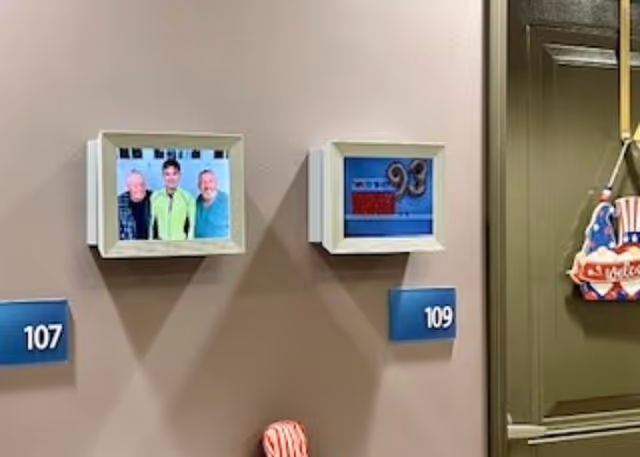 Two framed pictures mounted on a beige wall above blue room number plaques labeled 107 and 109. The picture above room 107 shows three people smiling, while the picture above room 109 shows a decorative item with the number 93. To the right, part of a green door is visible with a patriotic-themed hanging decoration.