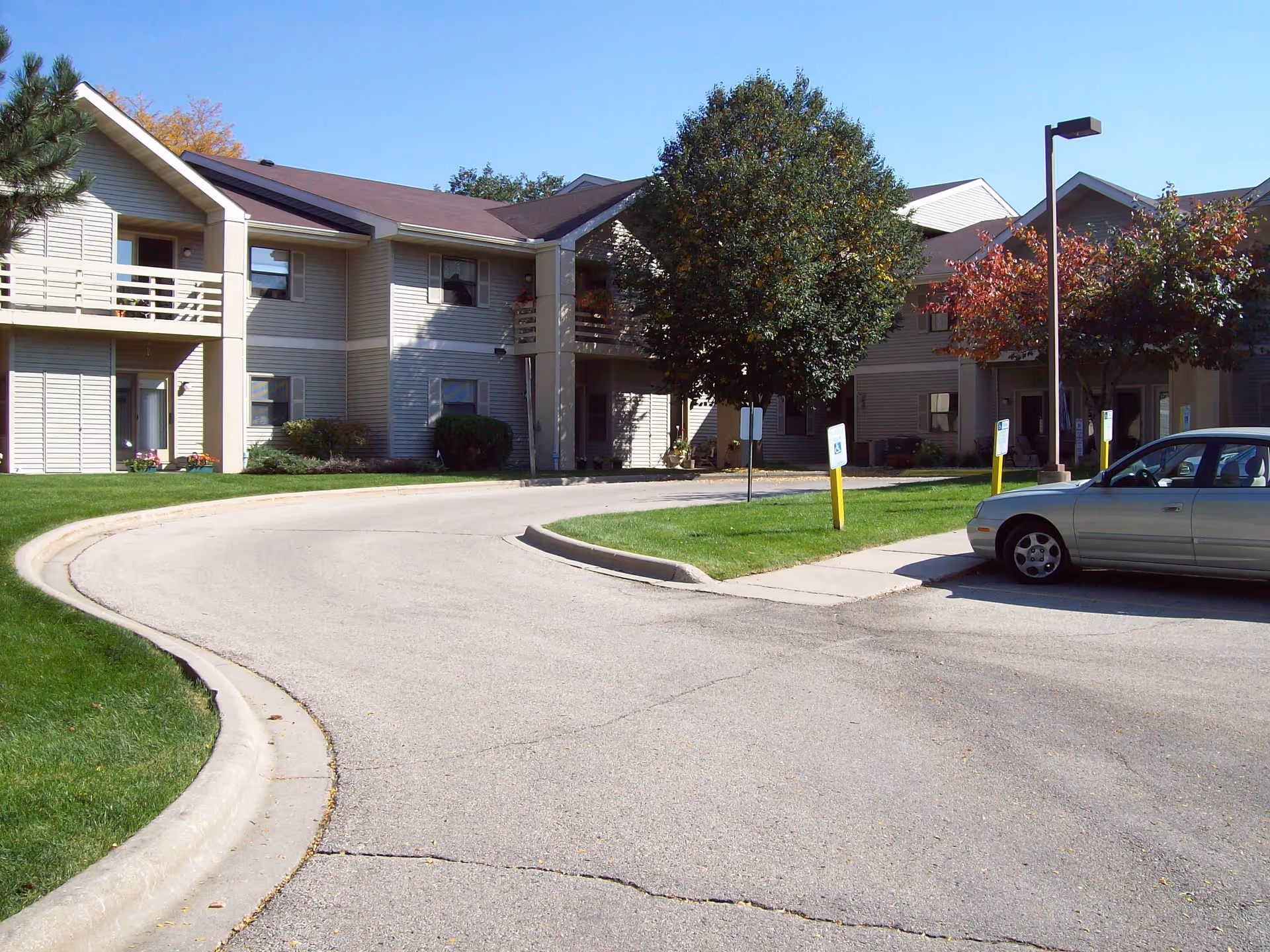 Exterior view of Meadow Grove Apartments showing a two-story residential building with balconies, surrounded by green lawns, trees, and a curved driveway with a parked silver car.